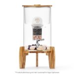 Ein Glas-Wasserfilter-Spender mit Holzdeckel steht auf einem dreibeinigen Ständer aus Akazienholz, im Inneren sind eine Filterkartusche und Edelsteine sichtbar.
